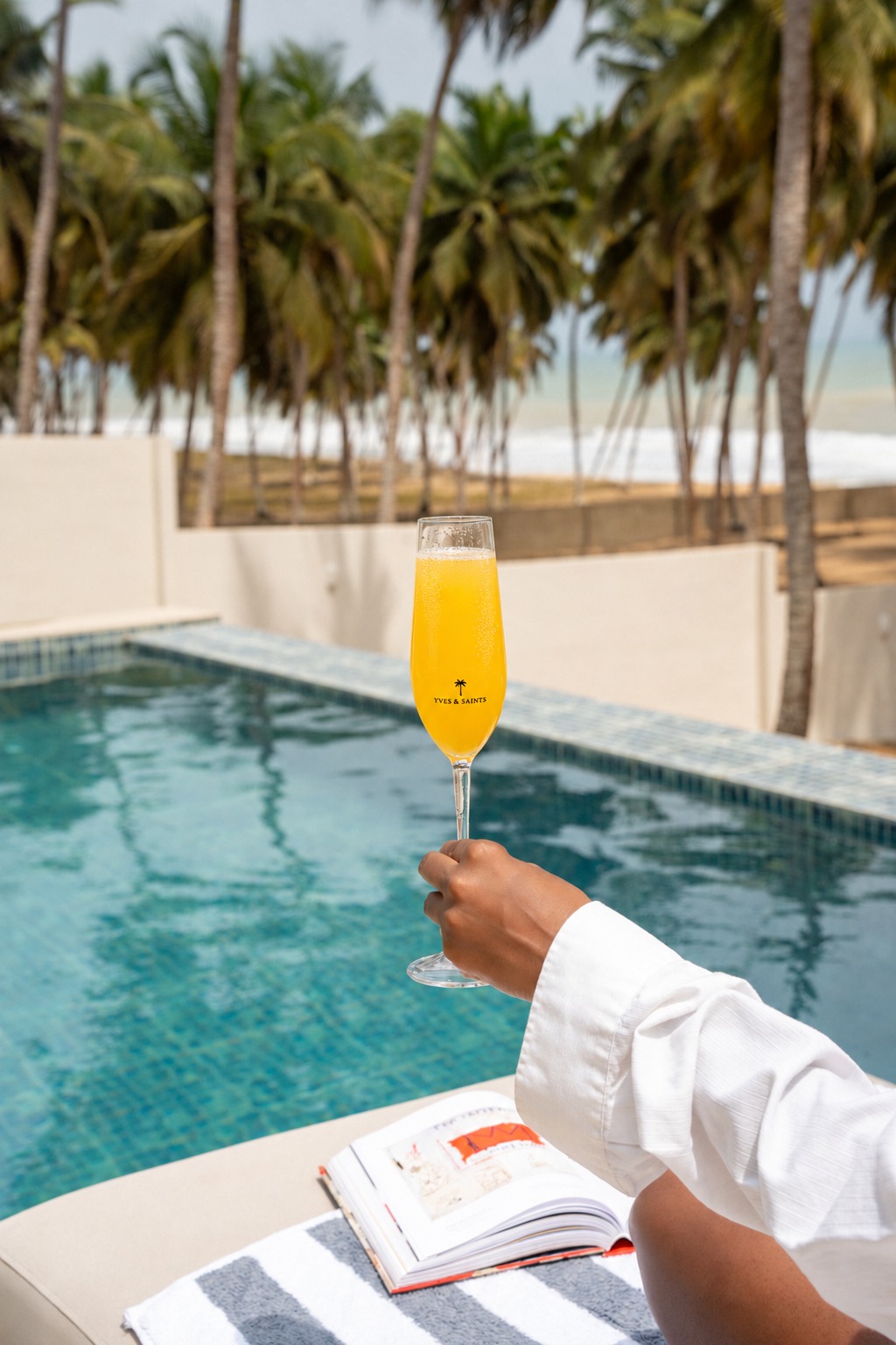 Guest raising a full orange mimosa in a branded Yves and Saints flute by the pool with palms and ocean beyond